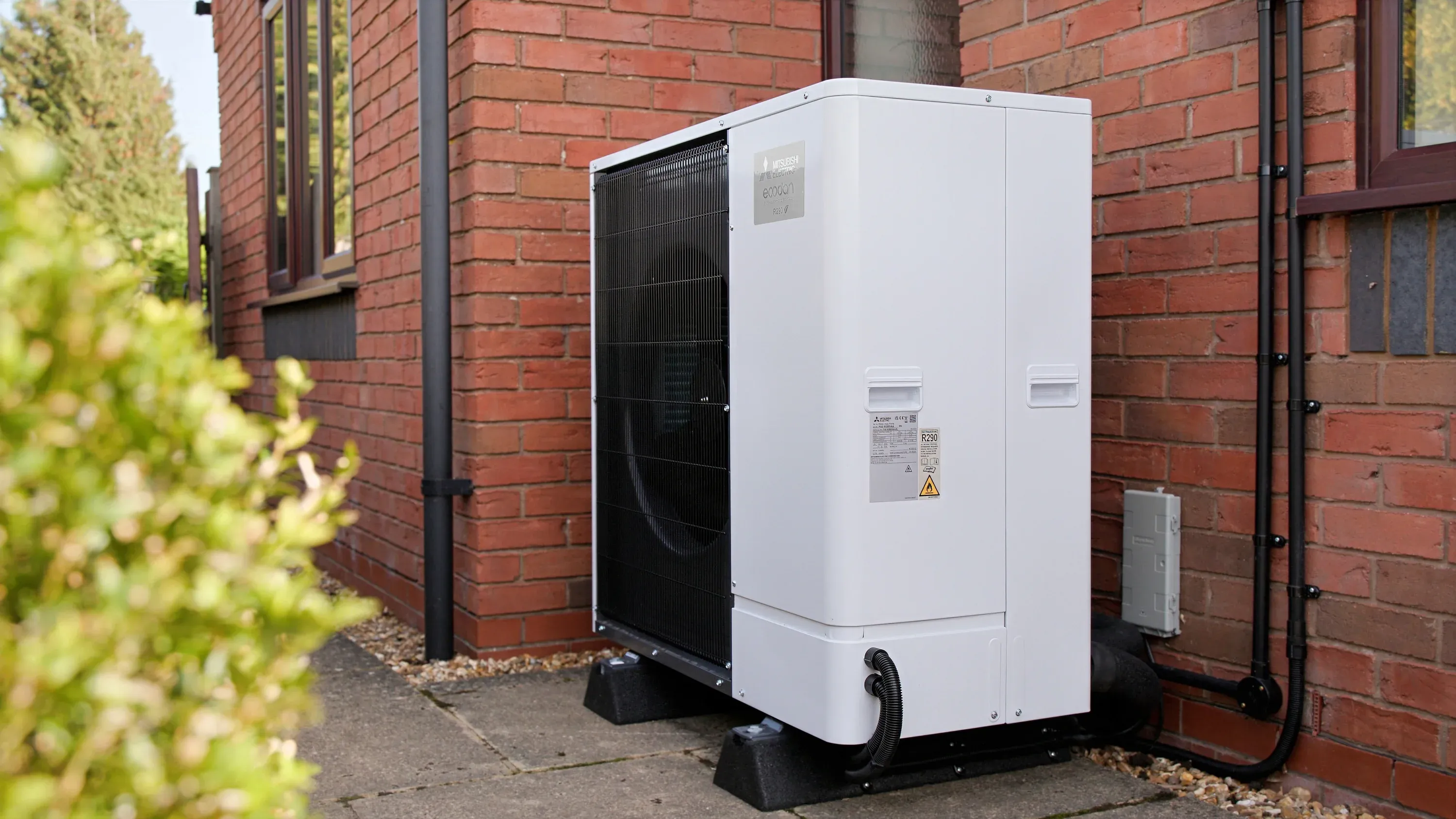 outdoor ecodan heat pump unit with white chassis and black front fan grill, installed in the corner of two exterior brick walls a brick home