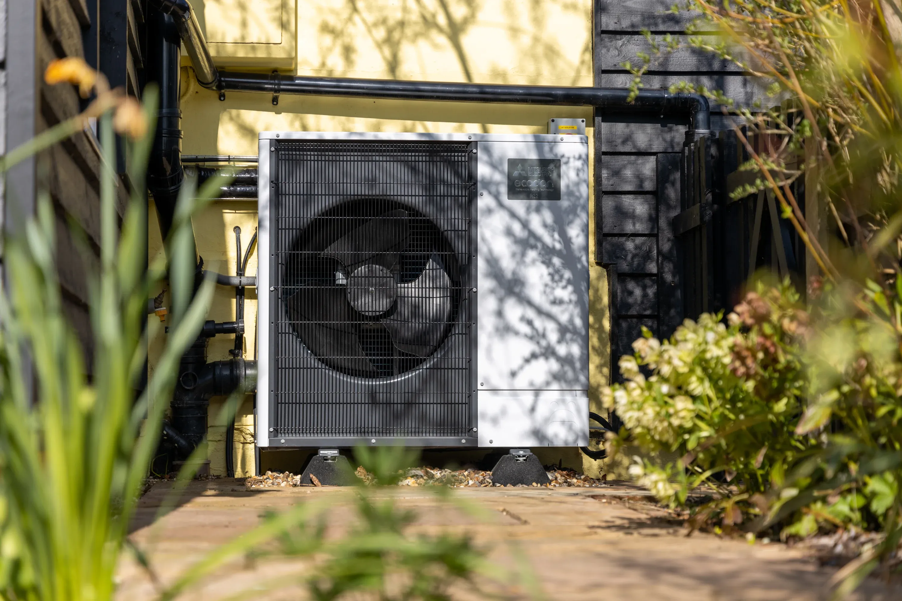 outdoor ecodan heat pump unit with white chassis and black front fan grill, installed alongside a home with  yellow and black exterior walls and decorative plants