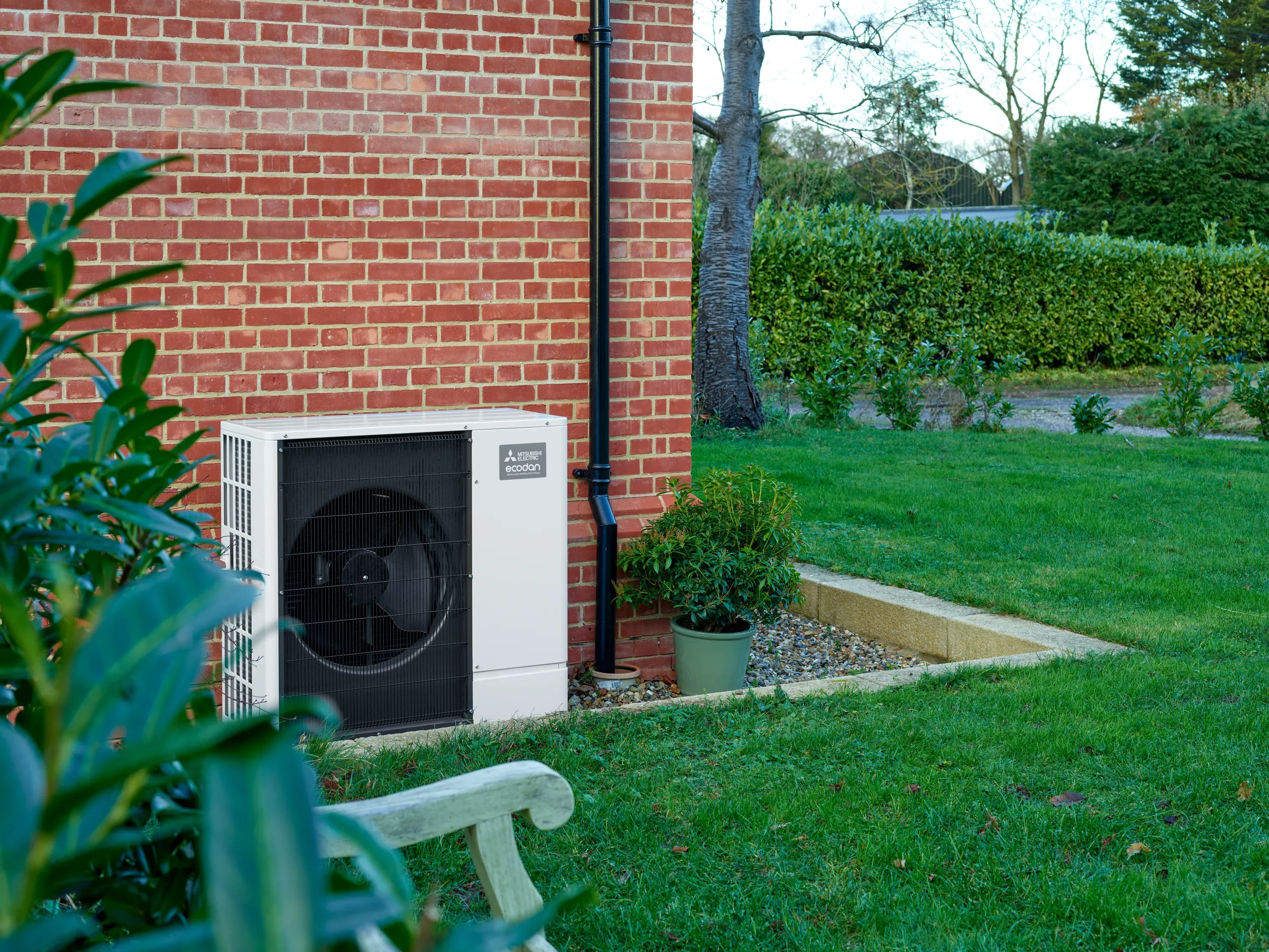 outdoor ecodan heat pump unit with white chassis and black front fan grill, installed alongside a brick home with decorative stone, plants, and emerald-green lawn. 