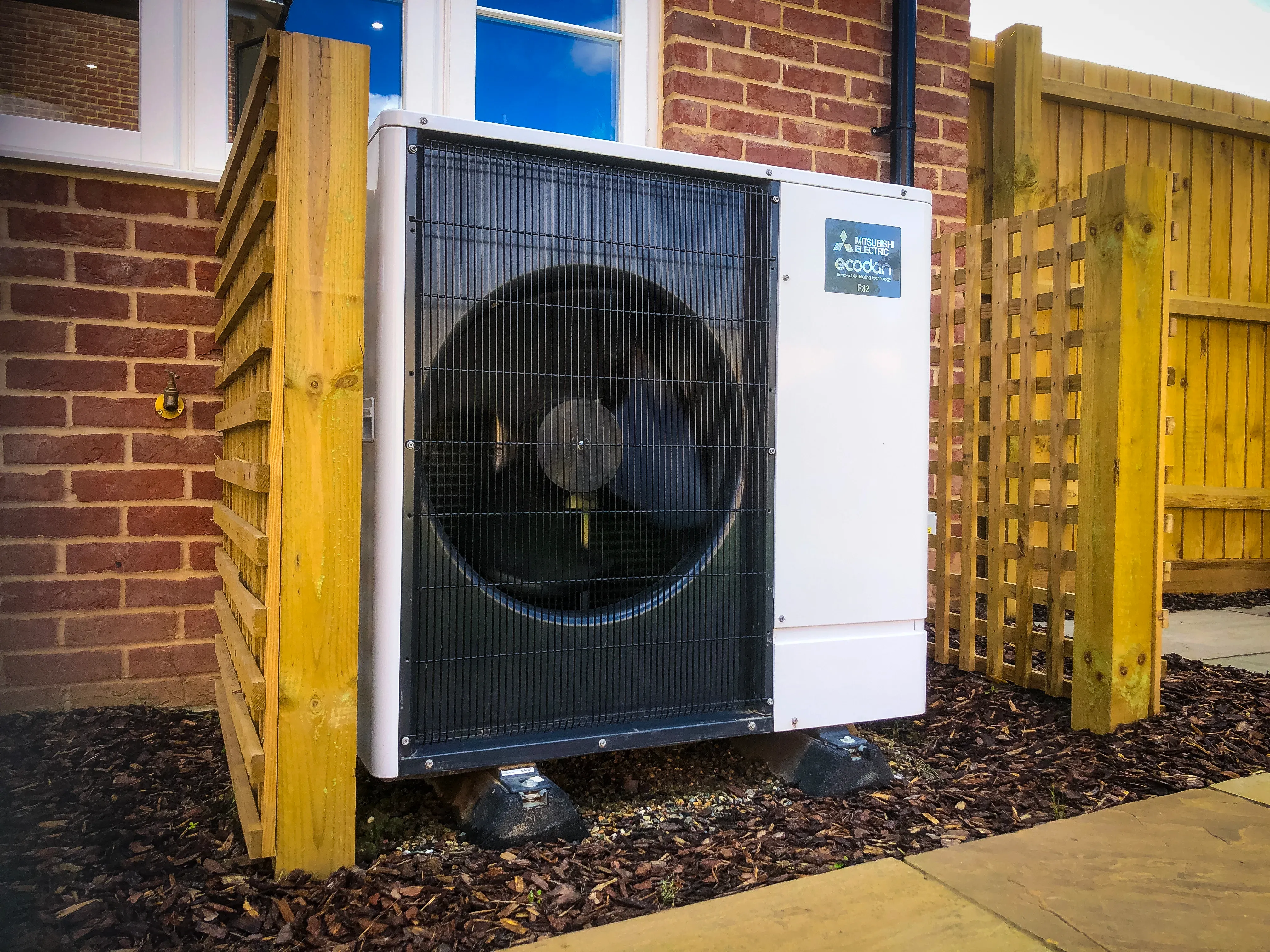 outdoor ecodan heat pump unit with white chassis and black front fan grill, installed alongside a brick home with decorative fencing